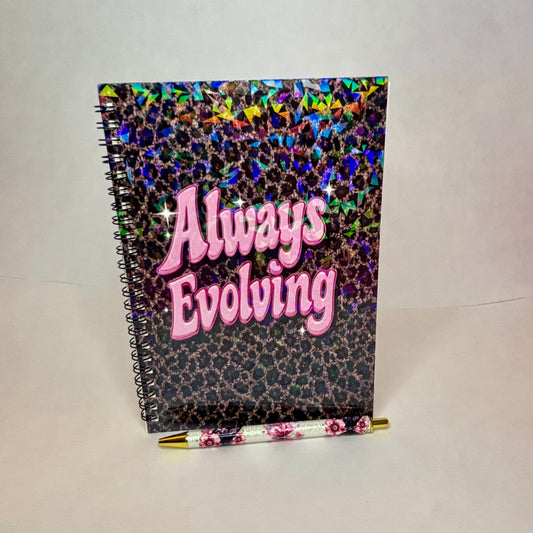 A spiral-bound notebook with a holographic leopard print cover and the words 'Always Evolving' in pink bubble letters, shown with a matching floral pen on a plain light background.