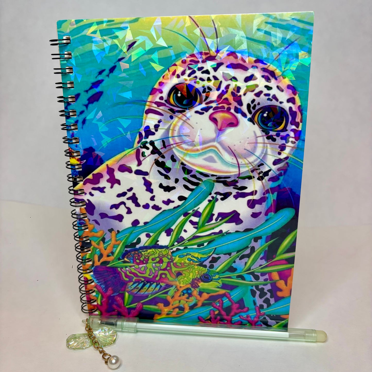 A spiral-bound notebook with a colorful, psychedelic seal painting on the cover, displayed with a clear plastic pen with a decorative charm.