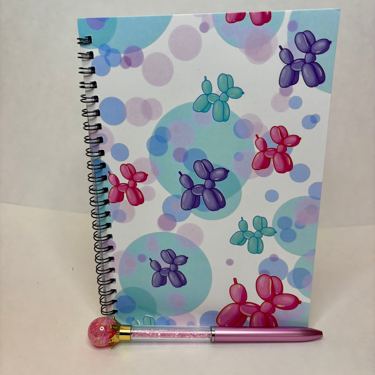 A spiral-bound notebook with a colorful balloon-dog pattern on a white background with pastel circles, standing upright next to a pink pen with a pink ball cap.