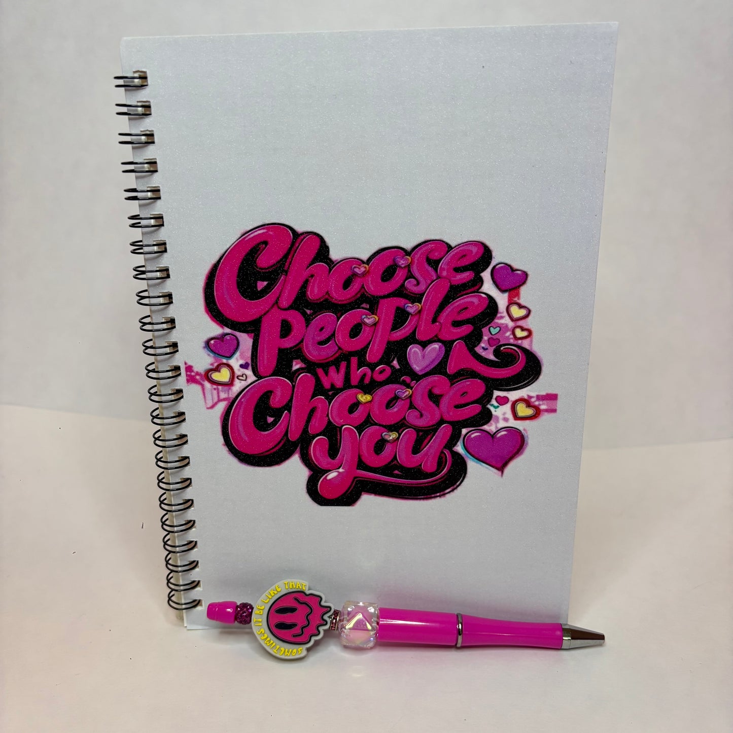 A white spiral-bound notebook with a bold pink and black graphic reading 'Choose People Who Choose You' decorated with hearts, shown with a matching pink pen on a light background.