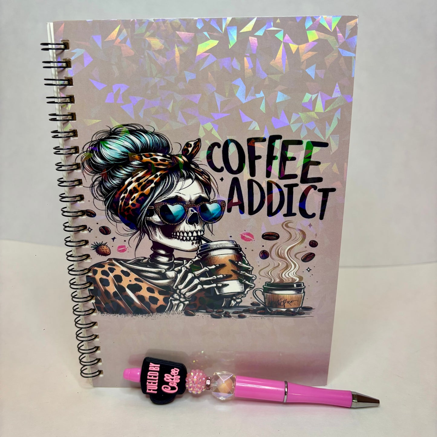 A spiral-bound notebook with a holographic cover featuring a cartoon skeleton coffee drinker and the words 'COFFEE ADDICT'. A pink pen with a 'FUELED BY Coffee' charm lies in front of the notebook on a white surface against a light gray background.