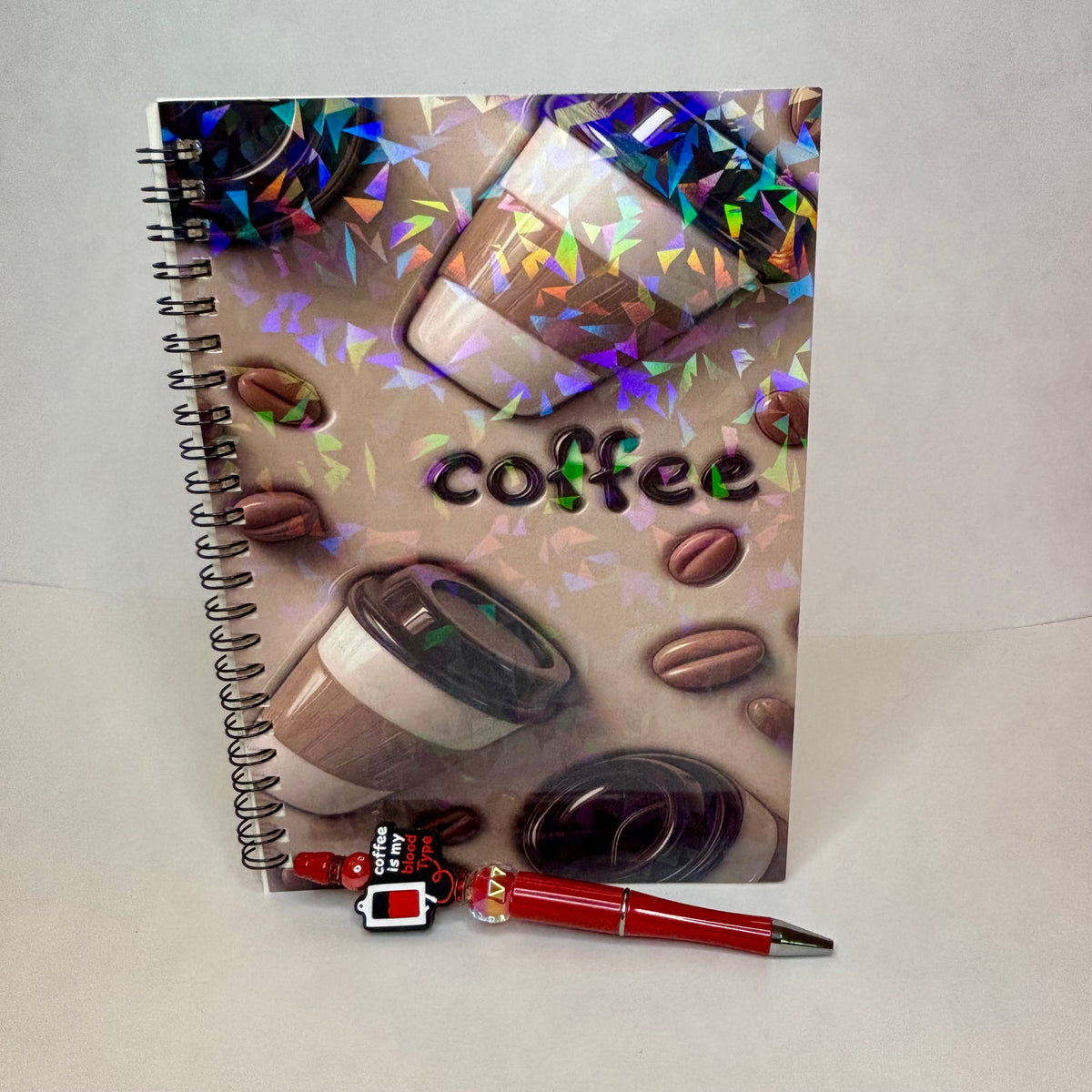 A spiral-bound notebook with a coffee-themed cover featuring coffee cups, coffee beans, and the word 'coffee' in holographic lettering, shown with a red pen in front.