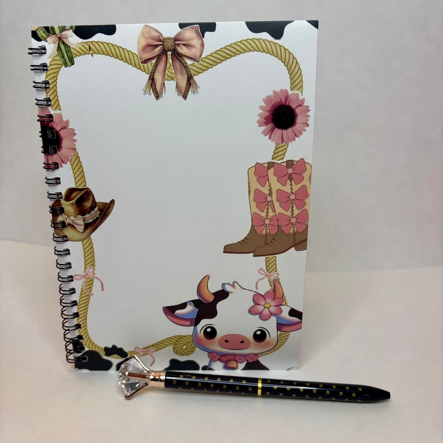 A spiral-bound notebook with a western cowgirl theme, featuring a cartoon cow, cowboy boots, a lasso, flowers, and a cowboy hat on a white cover, shown with a matching black pen that has a rhinestone handle.