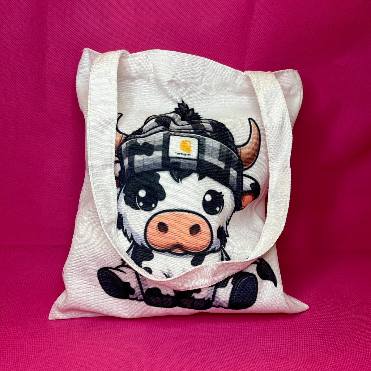 White canvas tote with a chunky cartoon black-and-white cow in a checkered beanie