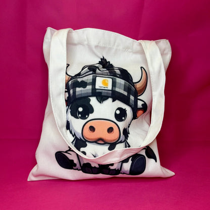 White canvas tote with a chunky cartoon black-and-white cow in a checkered beanie