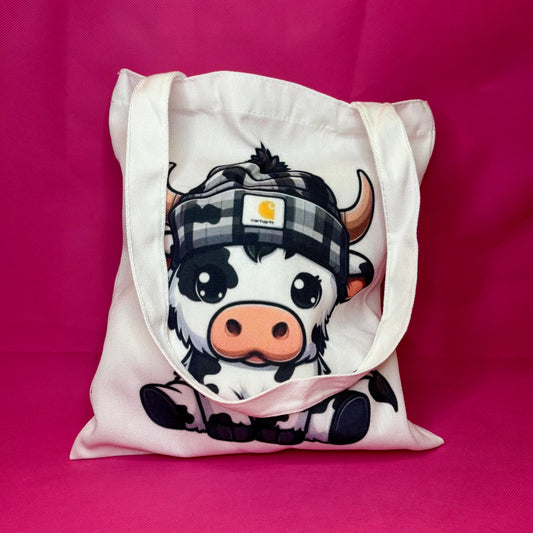 White canvas tote with a chunky cartoon black-and-white cow in a checkered beanie