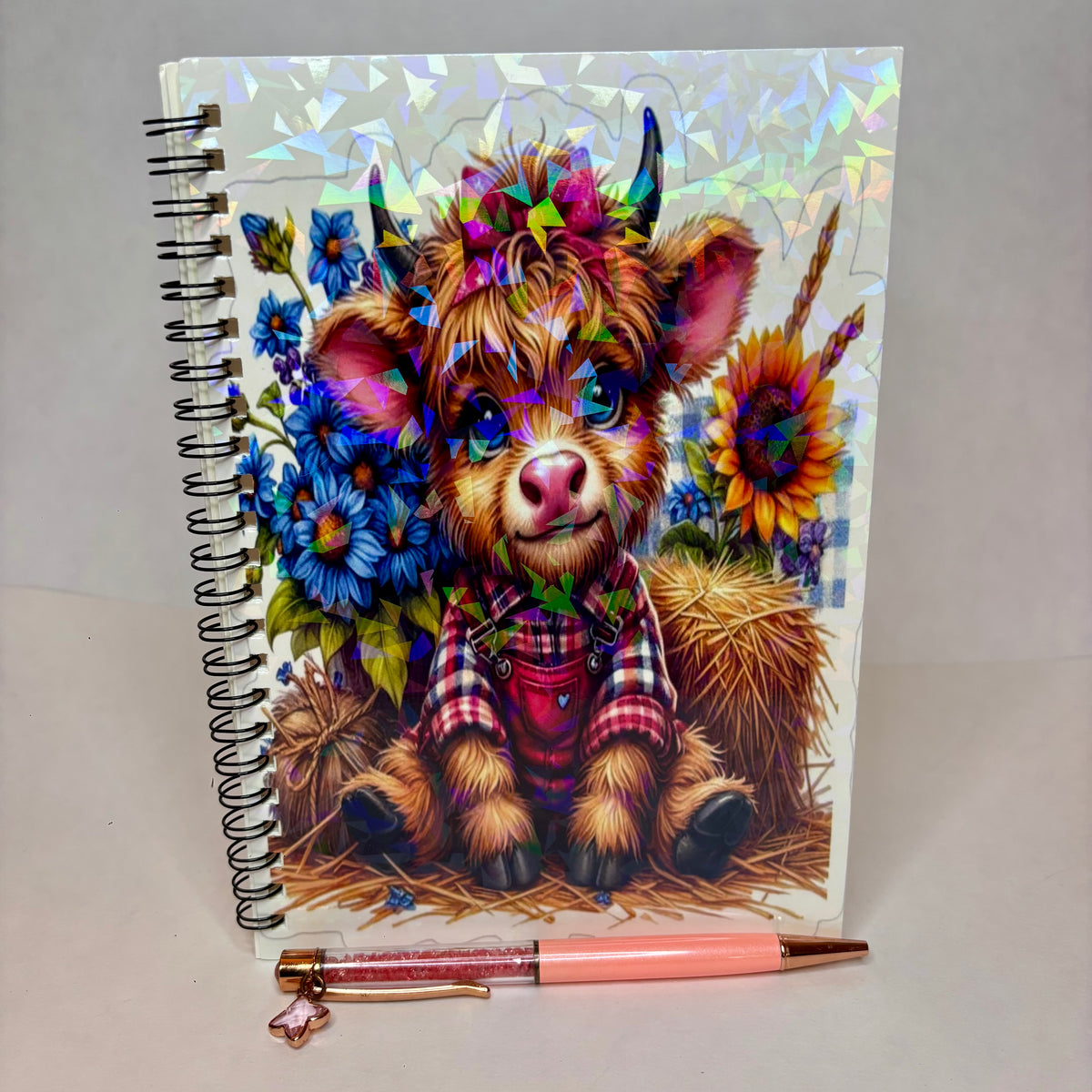 A spiral-bound notebook with a colorful illustration of a fluffy cow wearing a plaid shirt, surrounded by flowers and hay, shown on a light gray background with a pink pen placed in front.