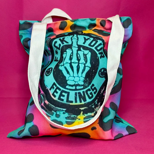 A canvas tote bag with a colorful tie-dye and leopard print pattern, featuring a large black graphic of a skeleton hand and the text 'B*CK YOUR FEELINGS' on the front. The bag has white shoulder straps and is photographed against a solid hot pink background.