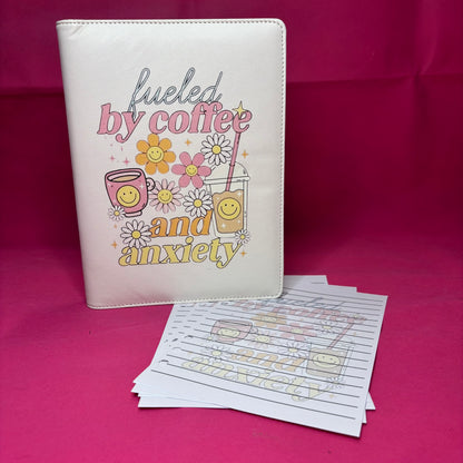 Cream A5 faux leather binder with 'fueled by coffee and anxiety' in pastel retro fonts, surrounded by smiley-faced latte, iced coffee, and daisies