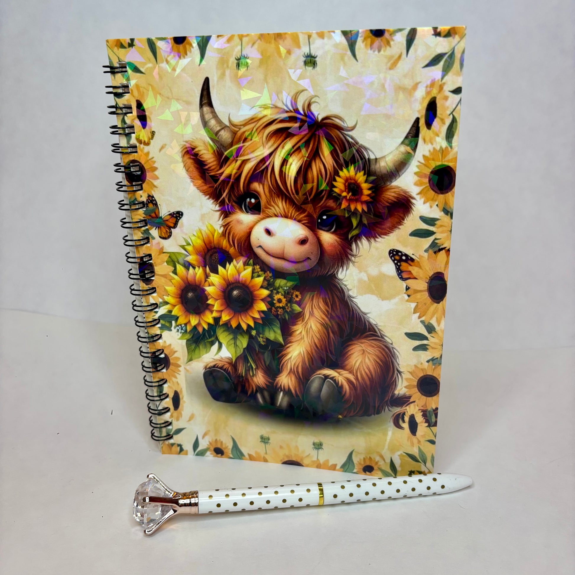 A spiral-bound notebook with a cartoon Highland cow surrounded by sunflowers and butterflies on a cream floral background, shown standing upright with a white pen with black polka dots and a rose gold cap placed in front on a white surface.