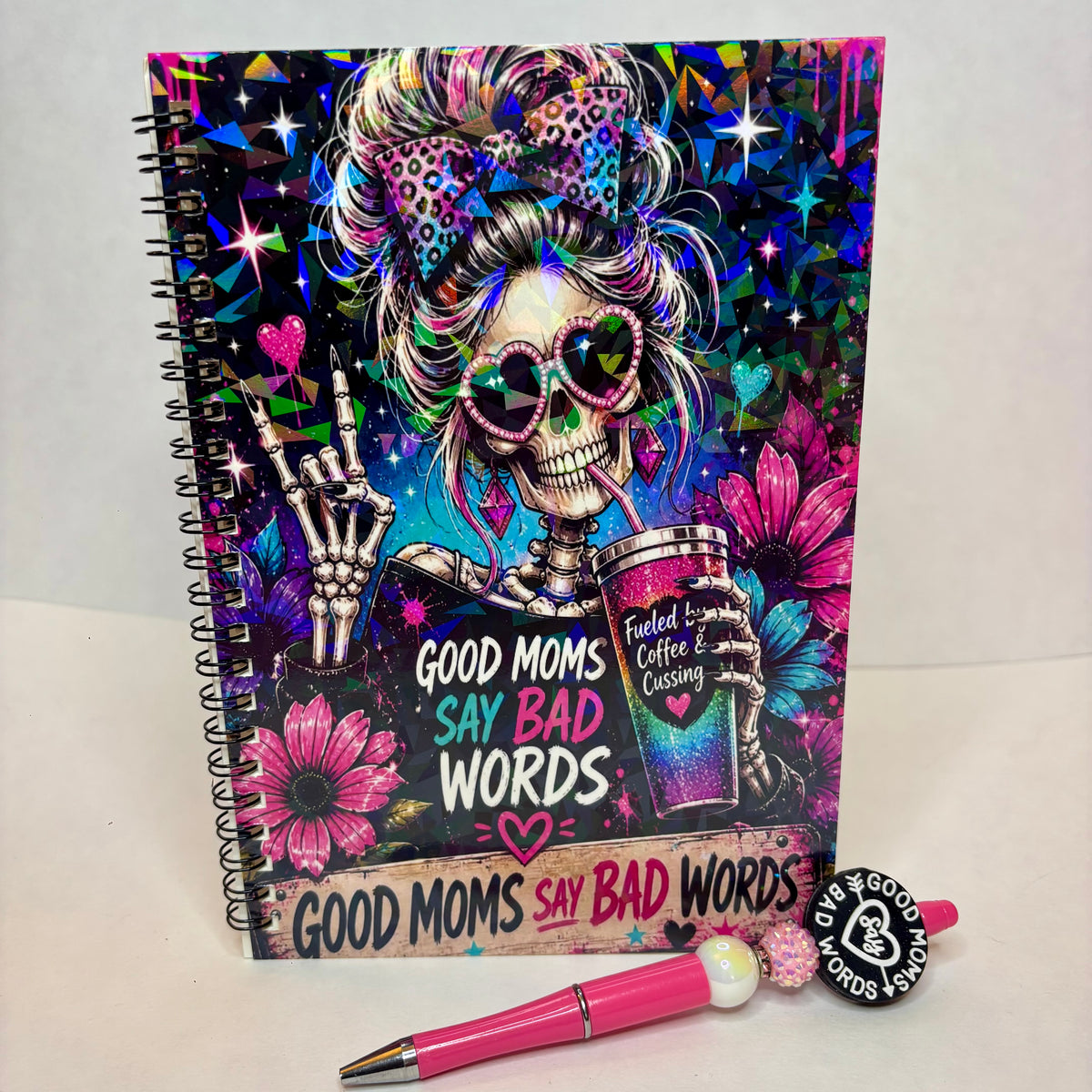 A spiral-bound notebook with a colorful Day of the Dead style skeleton illustration, shown standing upright on a white surface with a pink pen and a small matching charm in front.