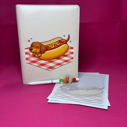 Cream A5 faux leather binder with a cartoon dachshund tucked inside a hot dog bun with mustard and ketchup on a red gingham picnic blanket