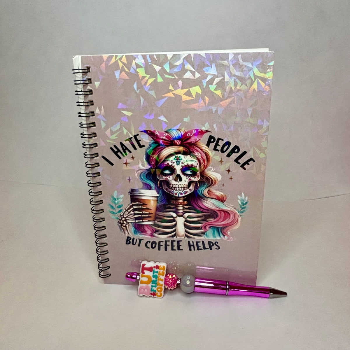 A spiral-bound notebook with a Day of the Dead sugar skull design and the text 'I HATE PEOPLE BUT COFFEE HELPS' on the cover, shown standing upright with a pink pen and small charm in front on a light gray background.