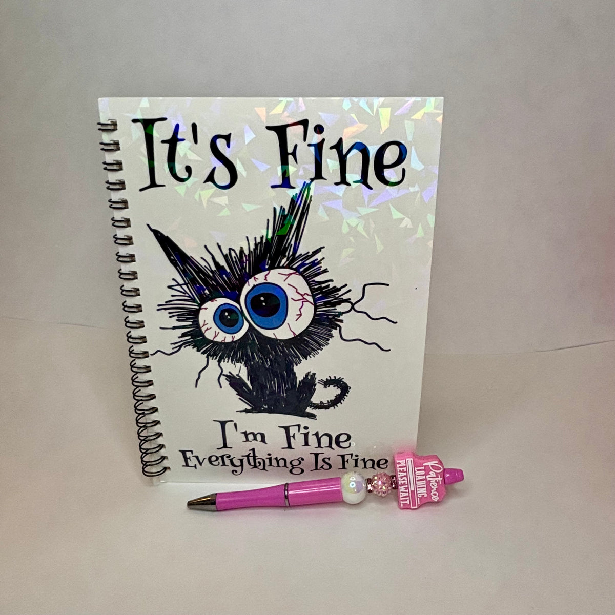 A spiral-bound notebook with a white cover featuring the text 'It's Fine' and 'I'm Fine Everything Is Fine' around a black spiky cartoon cat illustration. A pink pen and small pink charm are placed in front of the notebook on a plain light background.