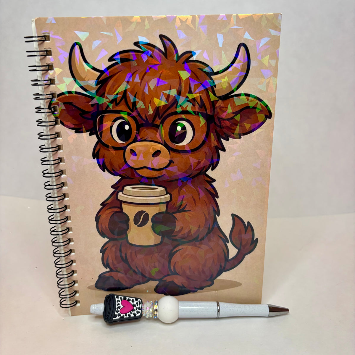 A spiral-bound notebook with a cartoon Highland cow character wearing glasses and holding a coffee cup, covered in holographic foil flakes. A white and black pen with a pink heart detail rests in front of the notebook on a plain light background.