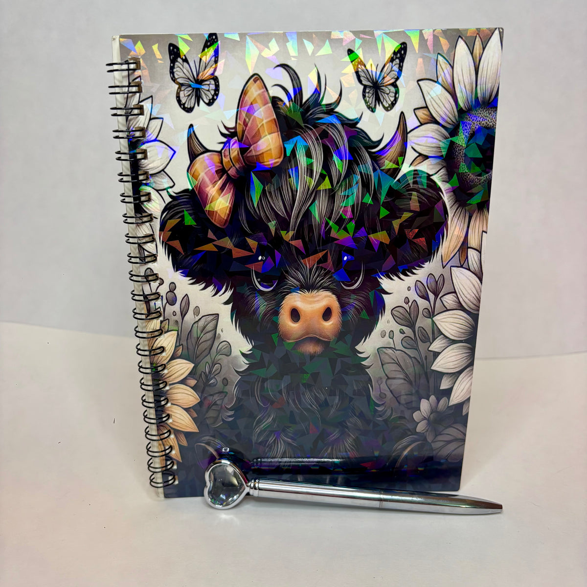 A spiral-bound notebook with a colorful, highly reflective holographic cover featuring a black fluffy animal face, butterflies, and sunflowers, shown with a silver pen resting in front on a white surface.