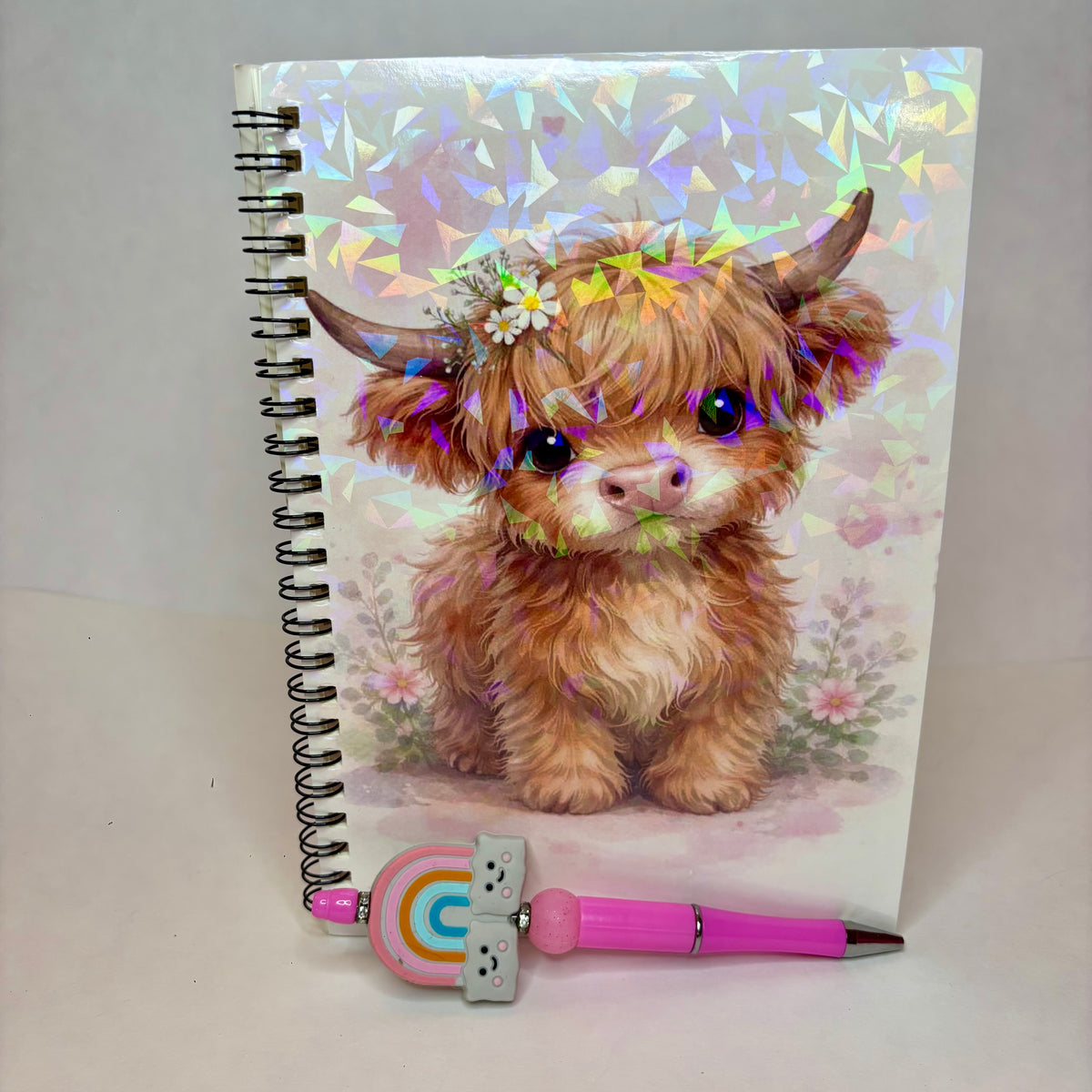 A spiral-bound notebook with a fluffy Highland cow illustration on the cover, shown standing upright with a pink pen resting in front. The notebook cover has a holographic, iridescent floral pattern, and the pen features a rainbow-colored bead.