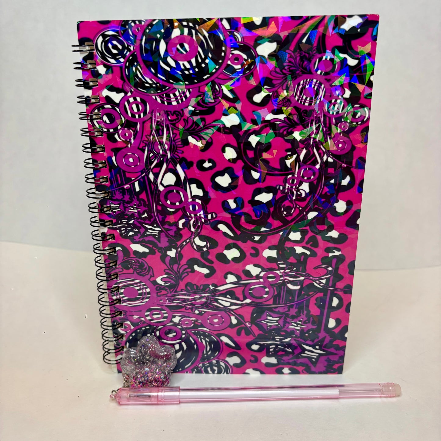 A spiral-bound notebook with a bright pink cover decorated with black leopard print and holographic silver glitter doodles, shown standing upright with a pink pen resting in front on a white surface.