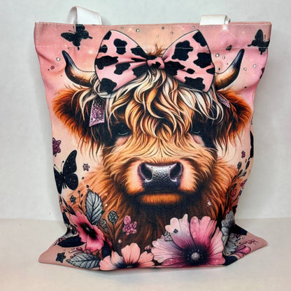 White canvas tote with a fluffy highland cow with cow-print bow on dusty pink background with black butterflies and pink florals
