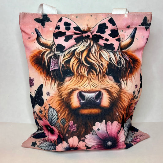 White canvas tote with a fluffy highland cow with cow-print bow on dusty pink background with black butterflies and pink florals