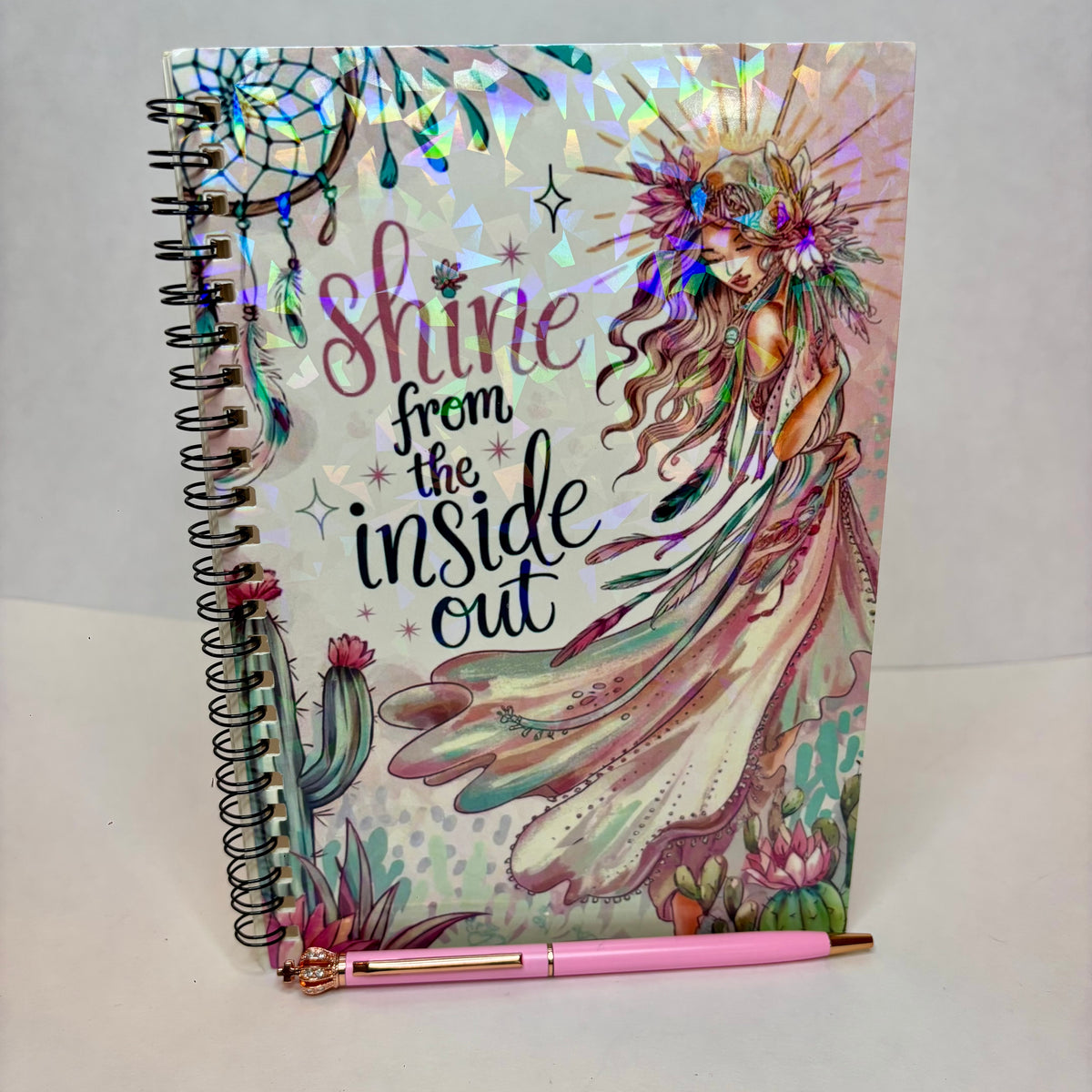 A spiral-bound notebook with a colorful fantasy illustration of a fairy and the phrase 'shine from the inside out' on the cover, shown standing upright with a pink pen resting across the bottom.
