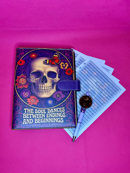 A purple journal with a skull and floral cover design, shown closed with a purple clasp. A pen and a stack of lined paper sheets are placed beside it on a bright pink background.