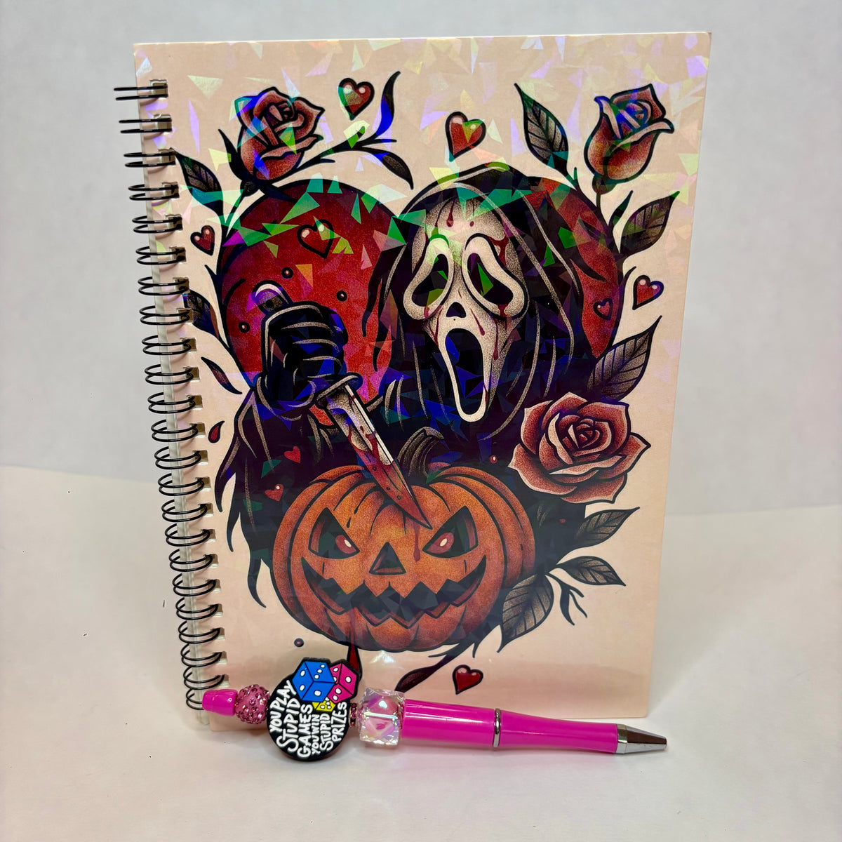 A spiral-bound notebook with a colorful Halloween-themed cover illustration featuring Ghost mask, a jack-o'-lantern, roses, and hearts on a holographic background. A pink pen with a decorative charm lies in front of the notebook on a white surface.